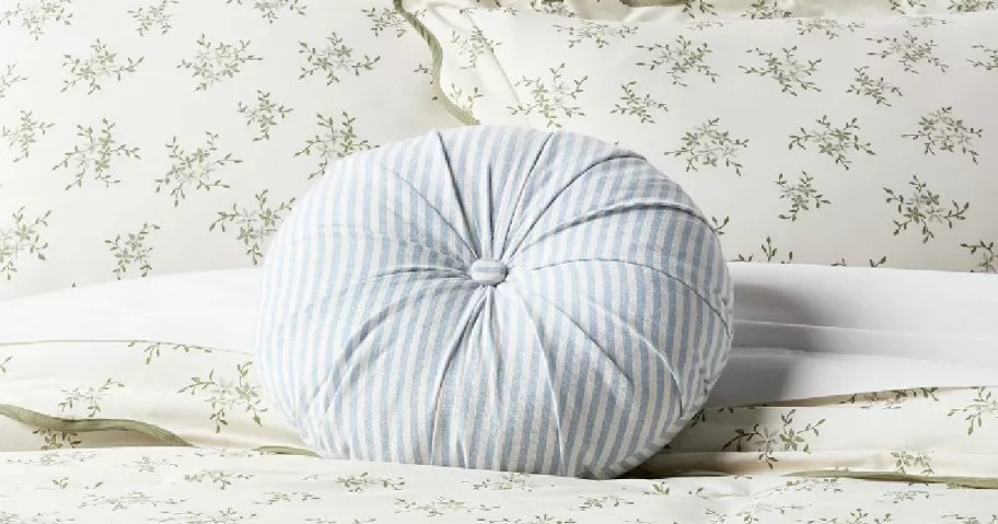 A round light blue and white striped tufted throw pillow on a floral bedset.