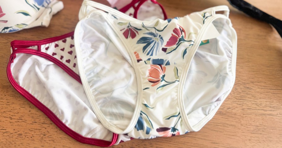 Two pairs of women's underwear resting on a wooden surface, with the top pair featuring a colorful floral pattern and the pair underneath displaying small hearts with red trim.
