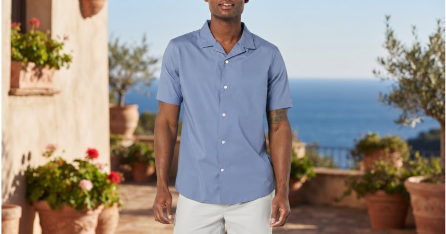 A man wearing a blue short-sleeve vacation shirt and white pants while standing on an outdoor patio with potted plants and an ocean view.