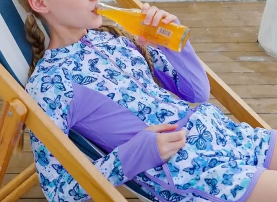 girl wearing a butterfly rash guard set while sitting in a beach chair and drinking an orange soda