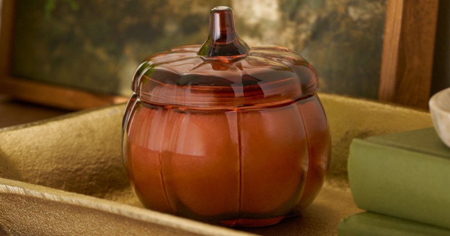A closed pumpkin-shaped jar candle sits on a gold tray next to a stack of green books.