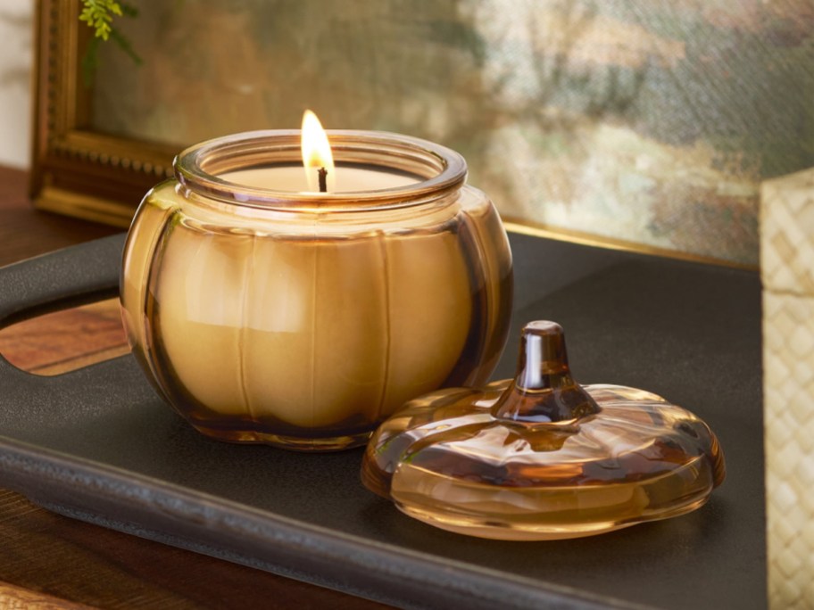 A pumpkin-shaped mason jar with a lit candle and lid sits on a dark tray.