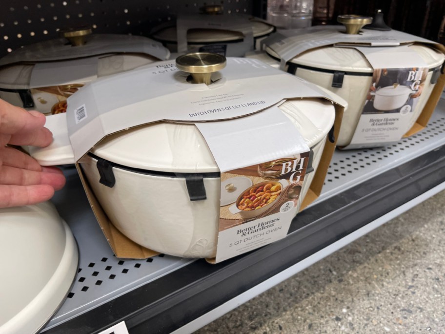 A hand reaching for the handle of a packaged, cream-colored Better Homes & Gardens 5-quart Dutch oven displayed on a gray store shelf next to identical items.