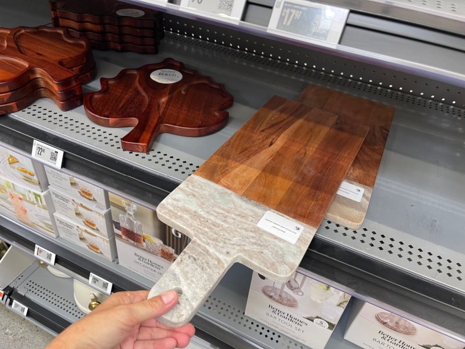 A hand holding a rectangular charcuterie board featuring a wood top and a light marble handle, displayed on a gray store shelf next to wooden leaf-shaped serving trays.