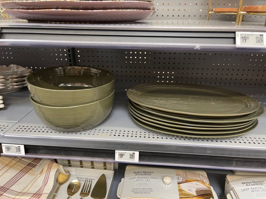 A stack of two olive green textured stoneware serving bowls and a stack of matching oval platters displayed side-by-side on a gray retail store shelf.