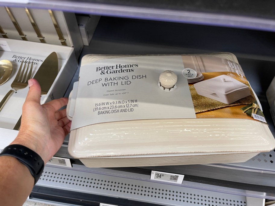 A hand reaching for a packaged, cream-colored Better Homes & Gardens deep baking dish with a lid displayed on a store shelf next to gold serving utensils.