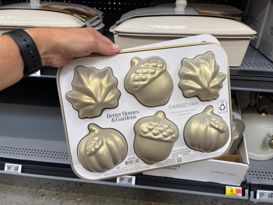 A hand holding a packaged Better Homes & Gardens gold cakelet pan featuring leaf, acorn, and pumpkin molds in a retail store aisle.