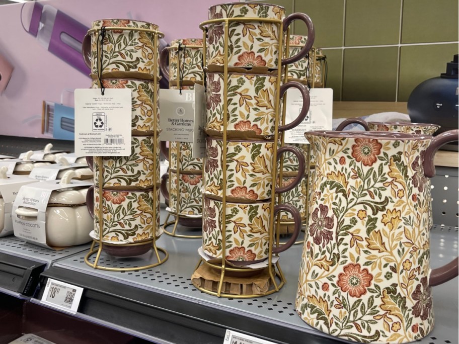 A store shelf displaying Better Homes & Gardens kitchenware, including sets of four stacking mugs in gold wire racks and a matching large pitcher, all featuring a vintage floral and leaf pattern in earthy tones.