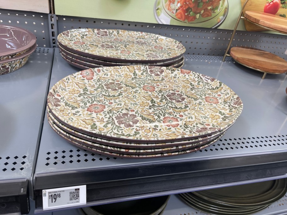A stack of large oval serving platters featuring an earthy vintage floral and vine pattern, displayed on a gray retail store shelf above a digital price tag.