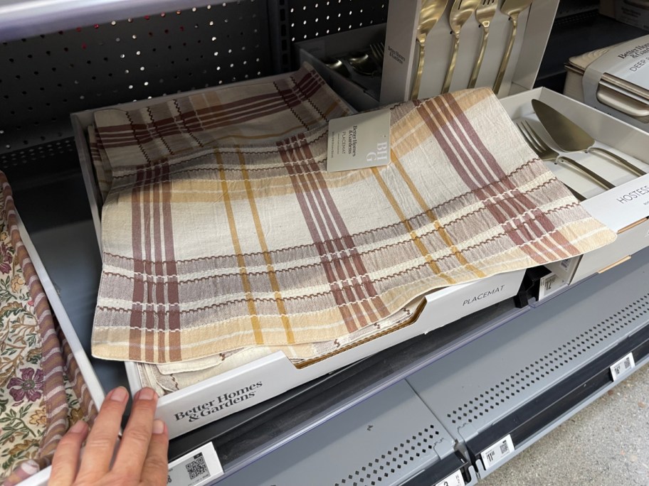 A stack of Better Homes & Gardens tan, brown, maroon, and yellow plaid placemats displayed in a white cardboard tray on a gray retail store shelf.