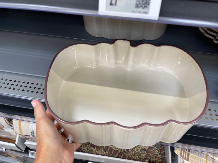 A hand holding a cream-colored, pumpkin-shaped ceramic baking dish with a dark rim on a store shelf.