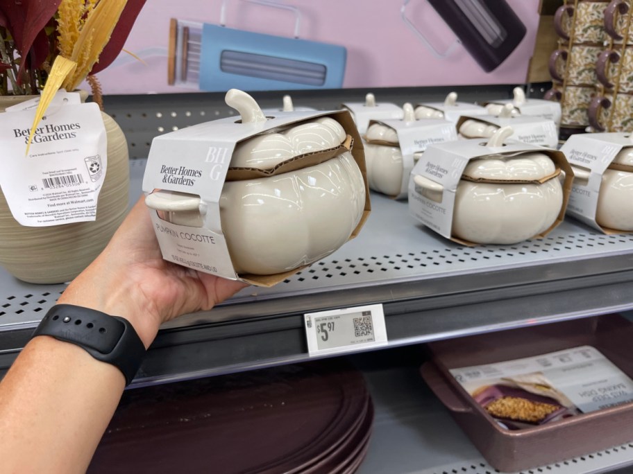 A hand holding a packaged Better Homes & Gardens white ceramic pumpkin cocotte with a lid, with several identical items displayed on a store shelf in the background.