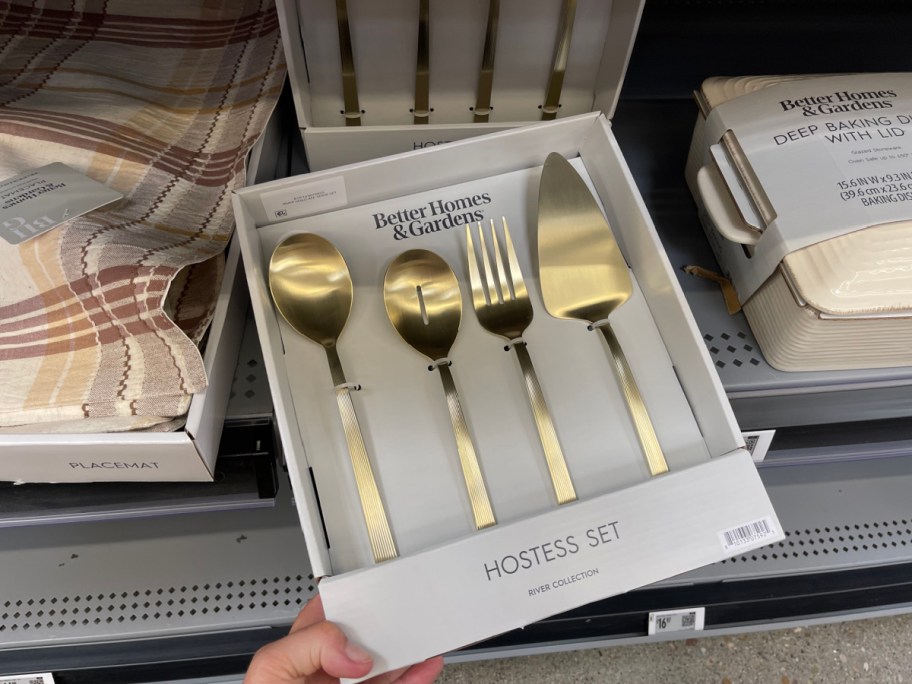A hand holding a packaged Better Homes & Gardens gold 4-piece hostess set featuring a serving spoon, slotted spoon, serving fork, and cake server, displayed on a gray retail store shelf.