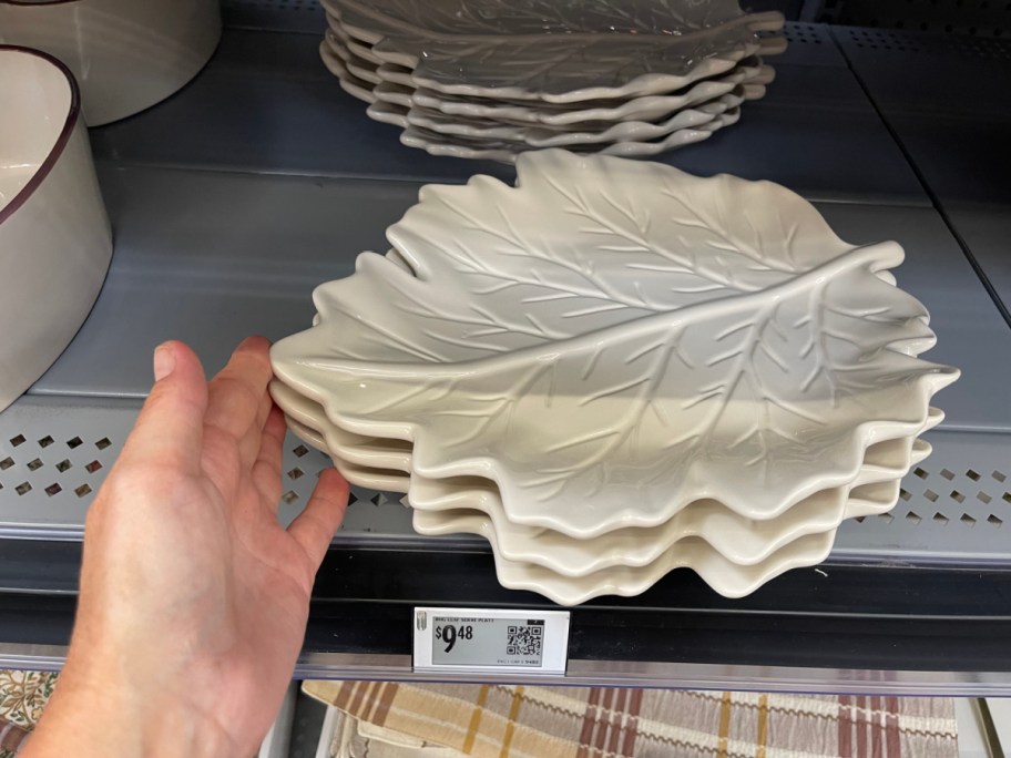 A hand reaching for a stack of sculpted, cream-colored ceramic leaf-shaped platters on a gray store shelf.