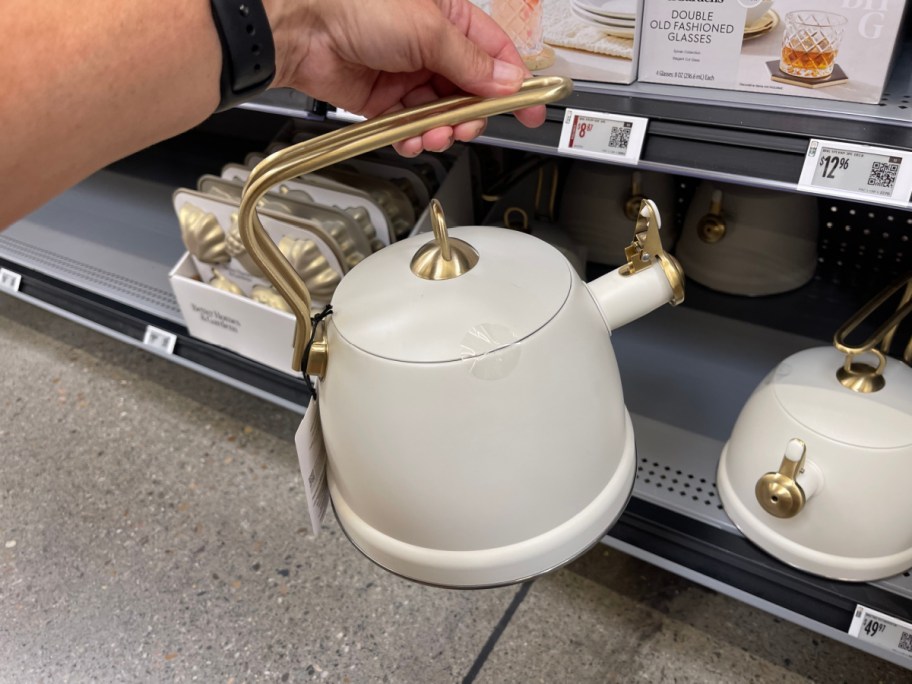 A hand holding a cream-colored Better Homes & Gardens whistling tea kettle with a gold handle and accents in a retail store aisle.