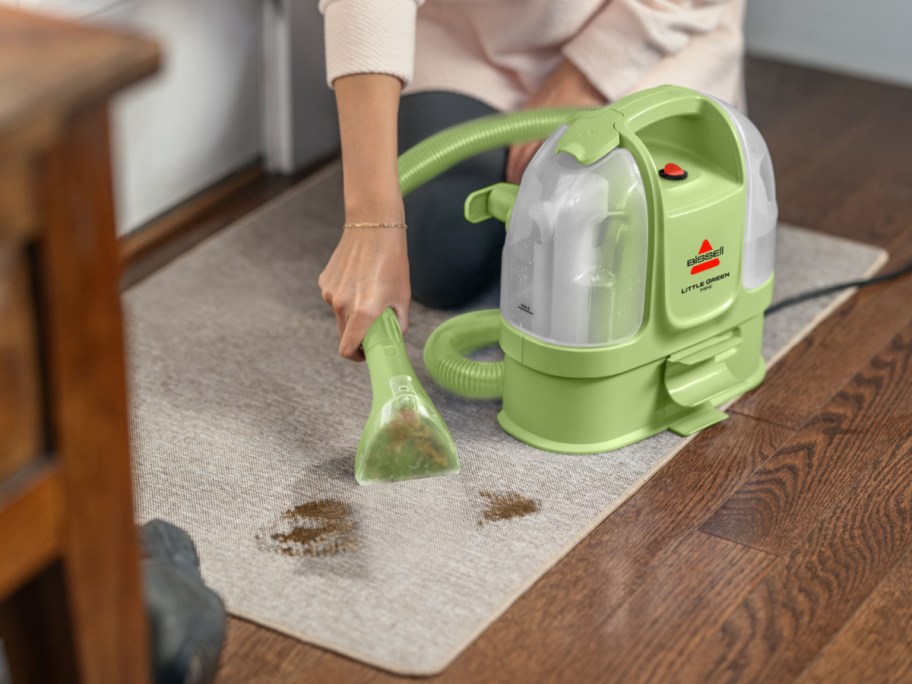 A person using a light green Bissell Little Green Mini cordless portable carpet cleaner to clean a spill on a rug.