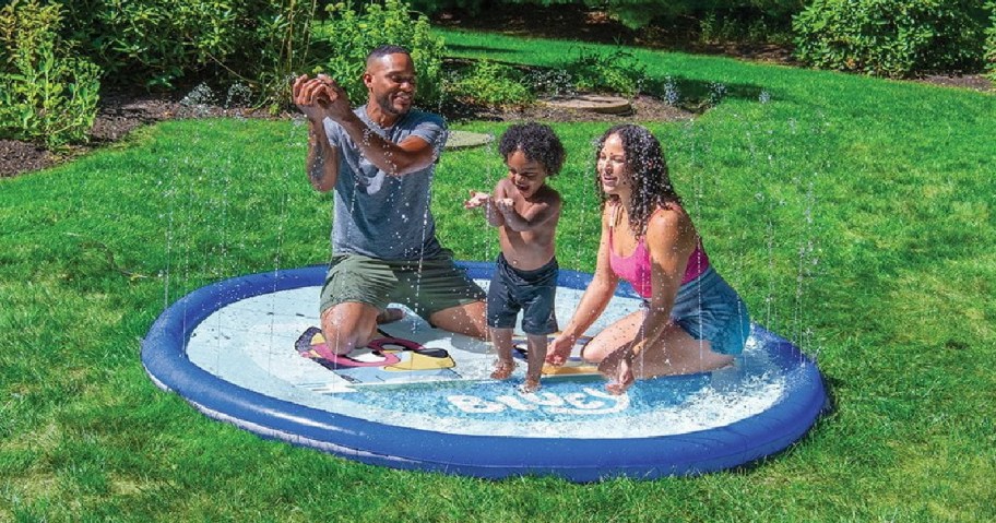 A family playing together in a blue-and-white Bluey-themed splash pad set up on a green lawn with water spraying upward from the edges.