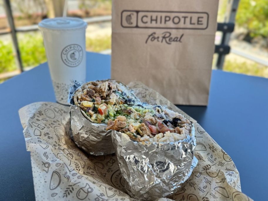 A chipotle burrito in a basket lined with paper in front of a peper cup with a straw and a paper Chipotle bag.