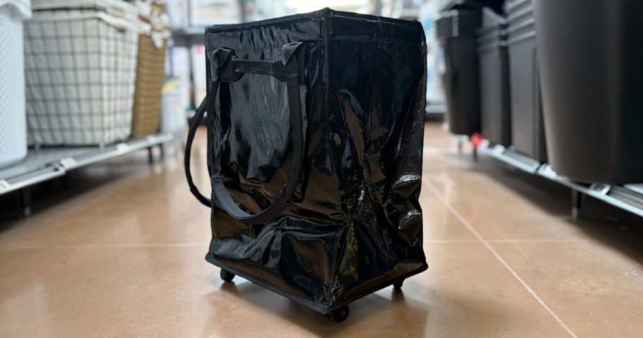 A black glossy rolling tote bag sitting on a tiled store floor in an aisle.