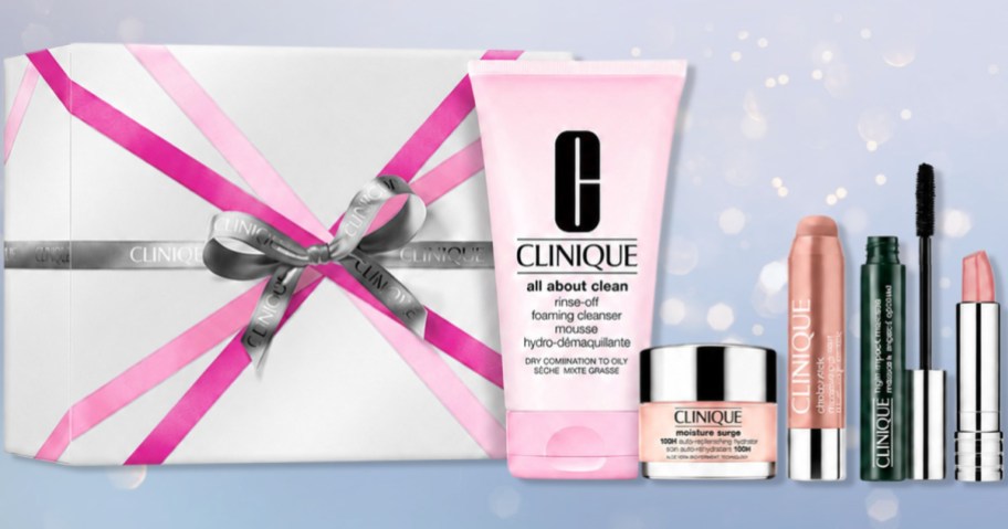 Clinique Ready To Glow Skincare and Makeup gift set featuring a foaming cleanser, moisture surge cream, chubby stick, mascara, and lipstick arranged in front of a wrapped white box with a pink bow.