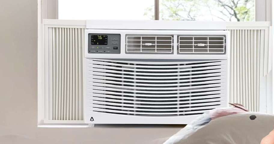 A white window air conditioner installed in a bright window with a digital display showing 76.