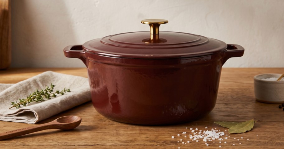 A dark red Dutch oven with a gold knob sitting on a wooden table next to a wooden spoon, fresh herbs, and scattered coarse salt.