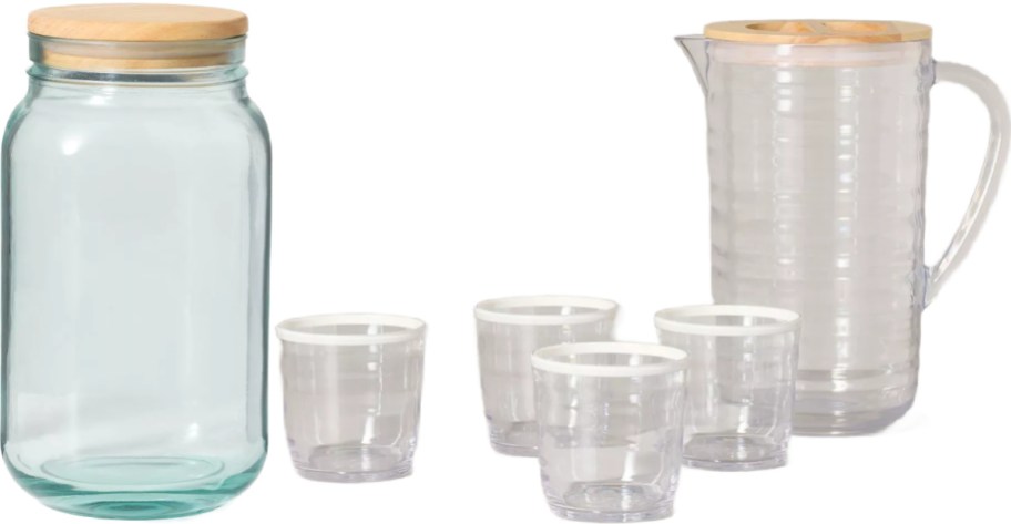 A clear glass canister with a wooden lid, four small clear cups with white rims, and a clear pitcher with a wooden lid against a white background.