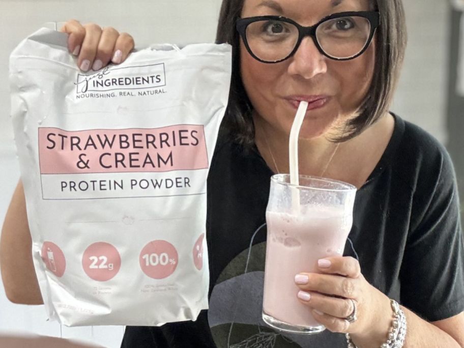Woman holding a bag of Just Ingredients Strawberry & Cream protein powder and drinking creamy pink liquid in front of a glass with a straw