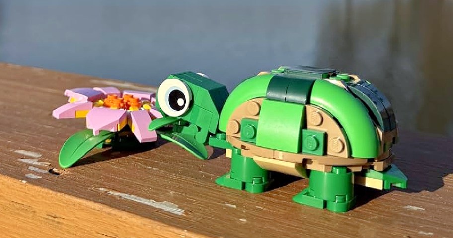 A small green and tan LEGO turtle standing next to a pink LEGO water lily on a wooden surface.