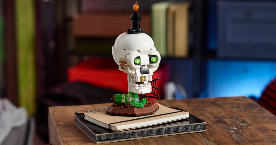A custom Lego Halloween skull model with glowing green eyes, a candle on top, and a spiderweb detail, sitting on top of notebooks on a wooden table.