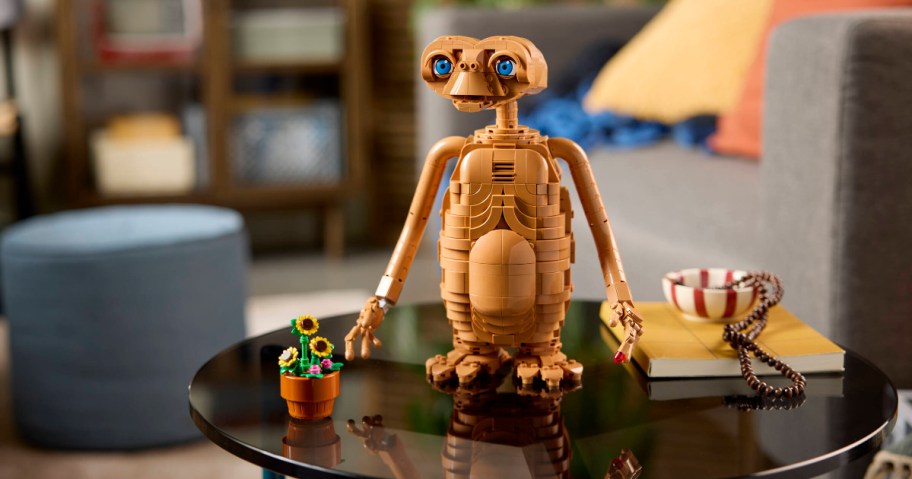 A LEGO E.T. the Extra-Terrestrial figure standing on a black glass table next to a small potted flower and a book with beads.