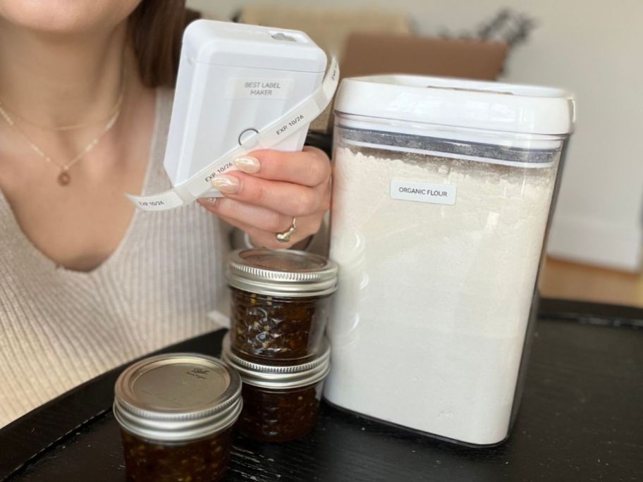 hand holding a NIIMBOT Wireless Label Maker next to a labeled container of organic flour and 3 small mazon jars of jelly