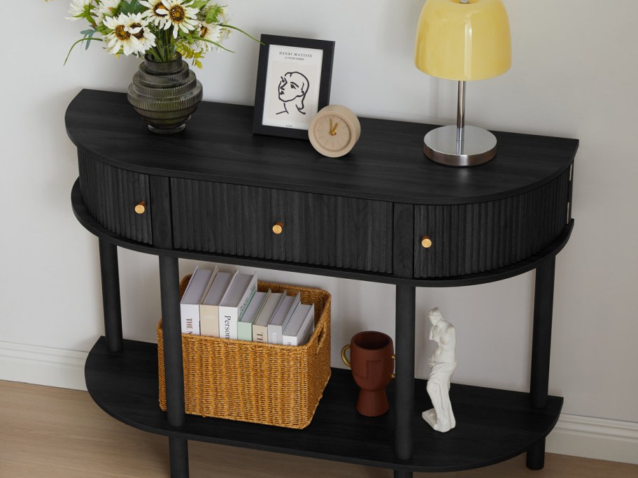 A black half-moon console table with fluted drawers and a lower shelf styled with a vase of flowers, a picture frame, a yellow lamp, a woven storage basket, books, and decorative objects against a white wall.