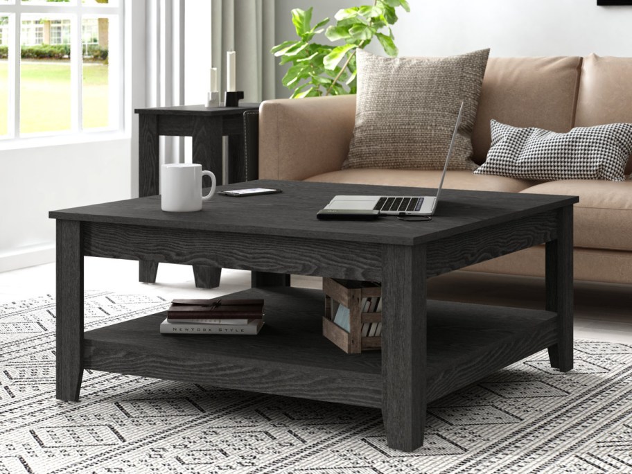 A square dark grey wooden coffee table with a lower storage shelf, styled with a laptop, mug, and phone on top, and books on the bottom shelf in a living room.