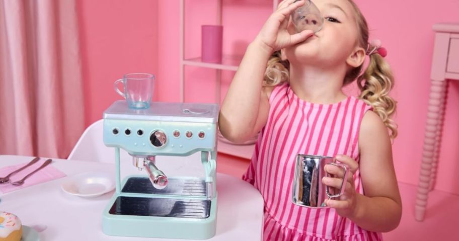 Little girl wearing a pink dress drinking out an espresso cup with a green little tikes espresso maker on the table