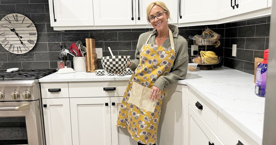 woman wearing a yellow floral print apron in kitchen and holding up a mixing bowl