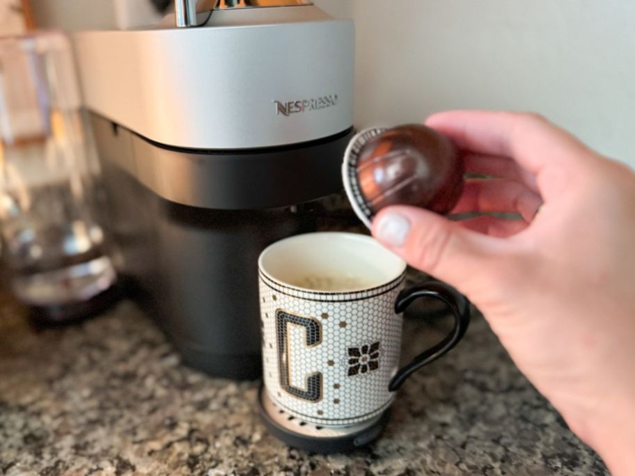 Hand holding a Nespresso Coffee Pod in front of an Anthropologie Tile Mug and a Nespresso Brewer