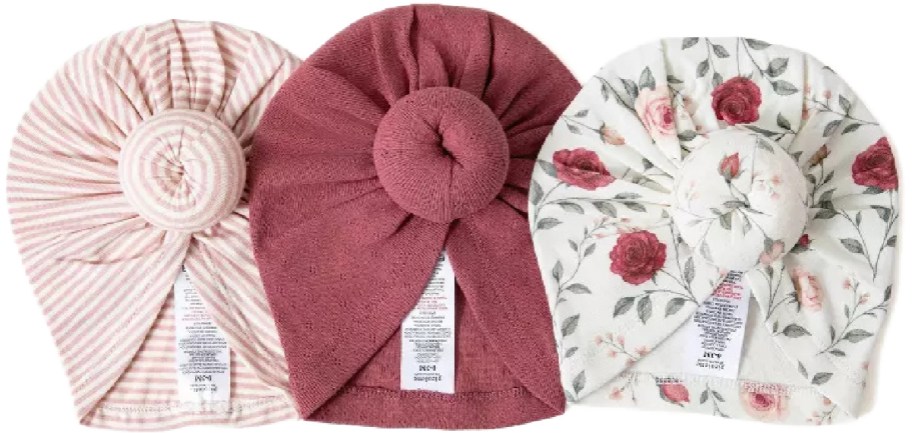 Three baby turban hats with top knots: one pink and white striped, one solid mauve, and one white with a pink floral pattern.