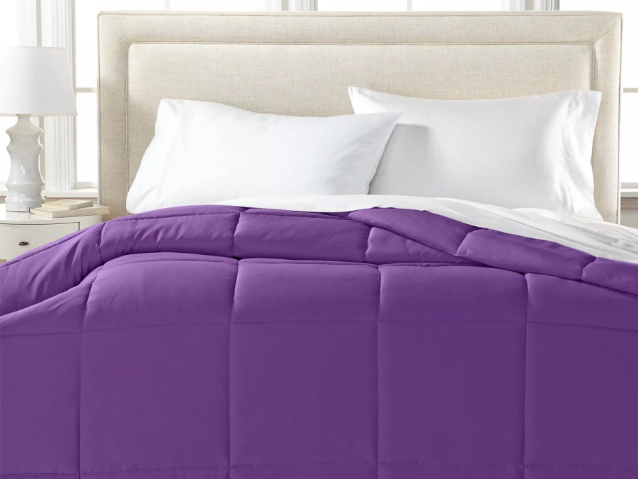 A Royal Luxe hypoallergenic down alternative comforter in a vibrant purple color, neatly made on a bed with white pillows and sheets against a padded headboard.
