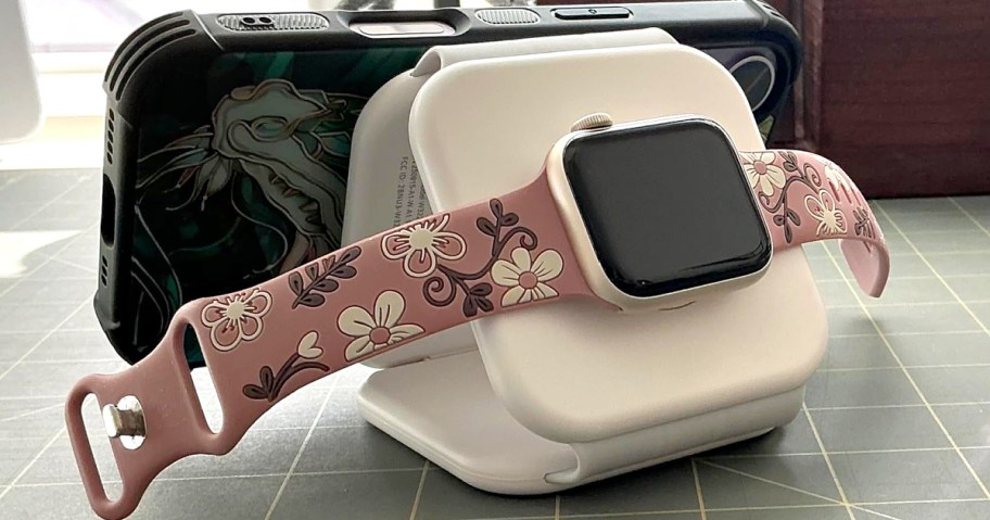 A white foldable charging station holding a smartphone in a dark green case and a smartwatch with a pink floral band.