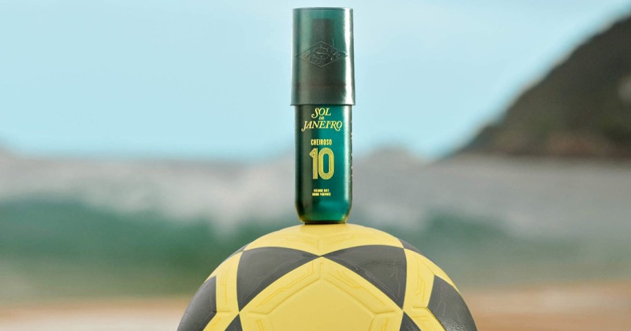 A dark green bottle of Sol de Janeiro Cheiroso 10 cologne mist resting on top of a yellow and black soccer ball against a blurred beach background.