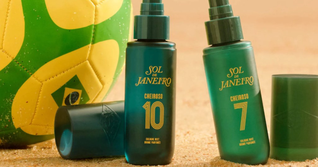 dark green bottles of Sol de Janeiro Cheiroso cologne mist resting next to yellow and black soccer ball against a blurred beach background.