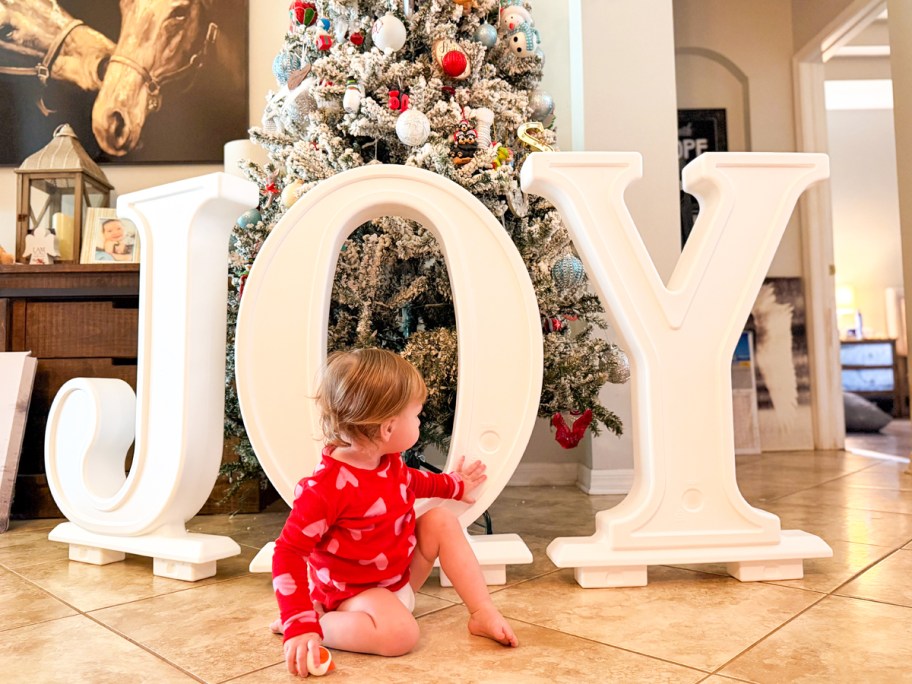Toddler on the floor in front of large white JOY letters with a Christmas tree in the background