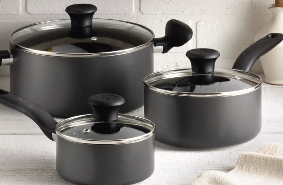 A T-Fal Initiatives aluminum nonstick cookware set featuring black pots and saucepans with glass lids and black handles.