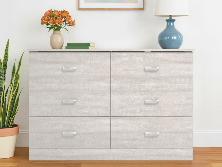 A light weathered grey Wade Logan Allyannah 6-drawer dresser styled with a vase of orange flowers, books, and a blue lamp on top against a white wall.