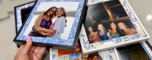 A hand holding up a Walgreens TilePix photo tile with a blue patterned border featuring two women hugging, with several other photo tiles displayed below.
