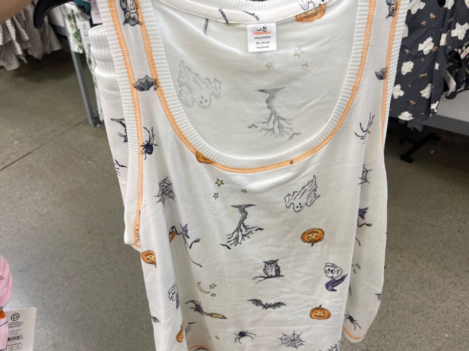 A white women's pajama tank top with orange trim hanging on a rack in a retail store, featuring a sketched Halloween pattern of ghosts, pumpkins, bats, owls, and spiders.