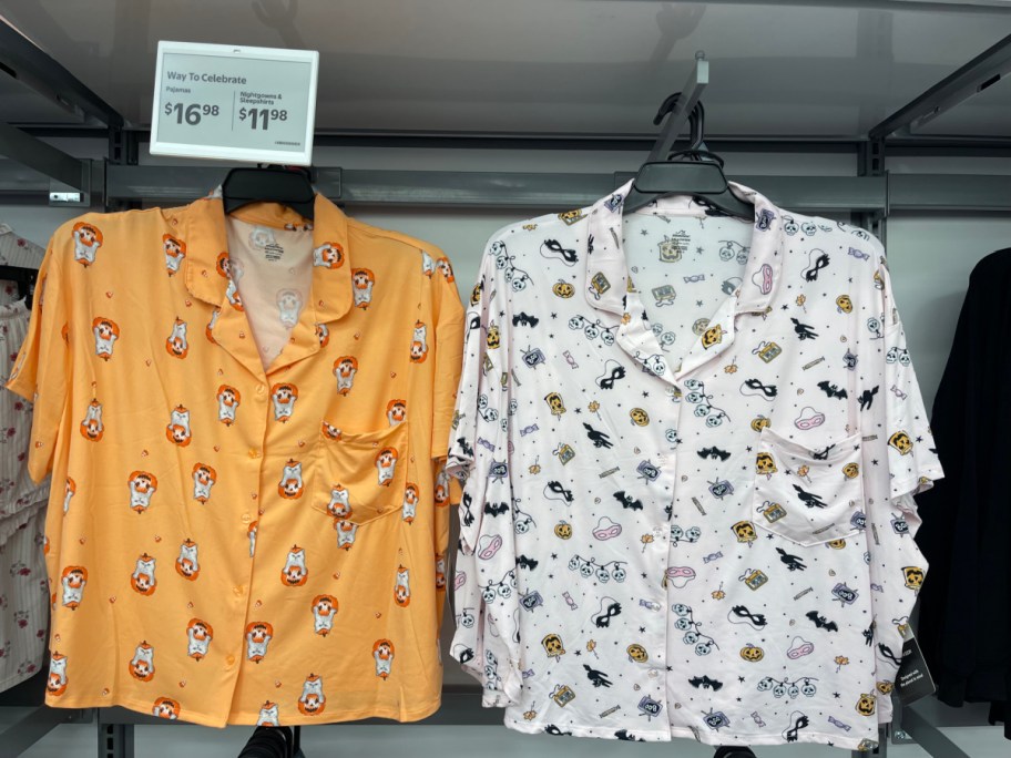 Two women's short-sleeve button-up Halloween pajama tops hanging on a retail clothing rack. The shirt on the left is orange with a cat and pumpkin pattern, and the shirt on the right is white with various Halloween motifs including bats, masks, and skulls.