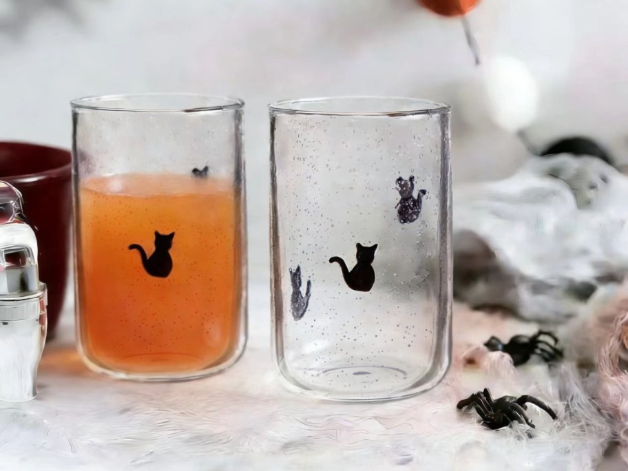 Two clear, bubbly drinking glasses decorated with black cats, one filled with an orange liquid, sitting among fake cobwebs and plastic black spiders.