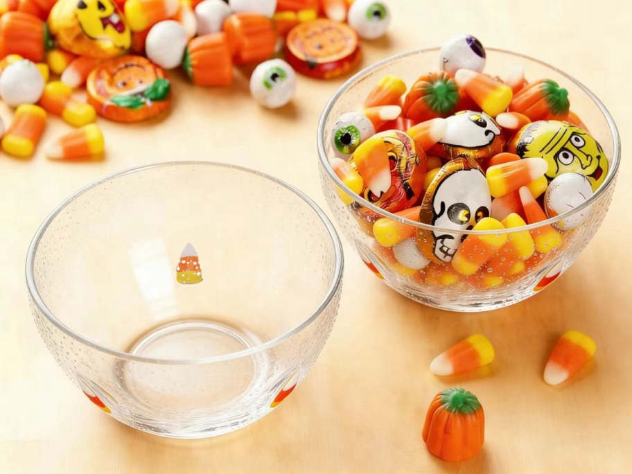 Two clear, bubbly glass bowls decorated with candy corn, one empty and one filled with assorted Halloween candy, sitting on a wooden surface with more candy scattered around.